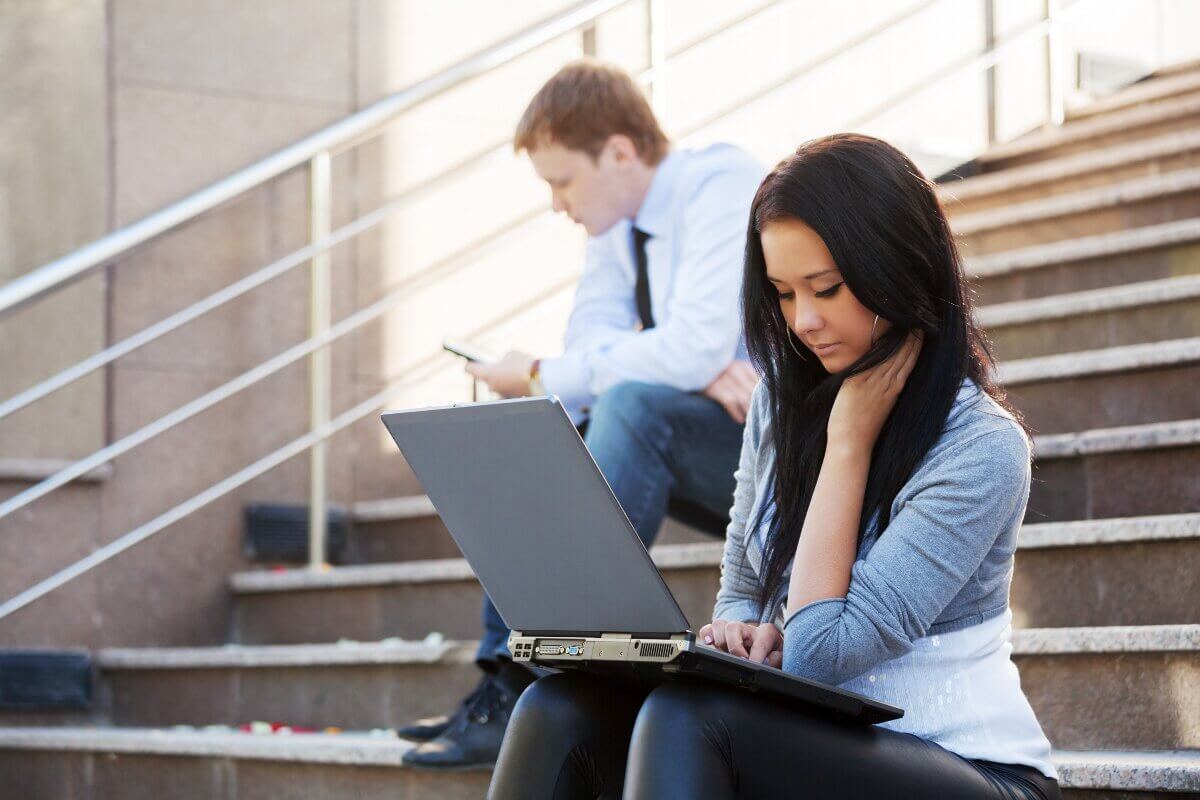 Eine junge Frau sitzt mit Laptop auf den Knien auf einer Außentreppe.